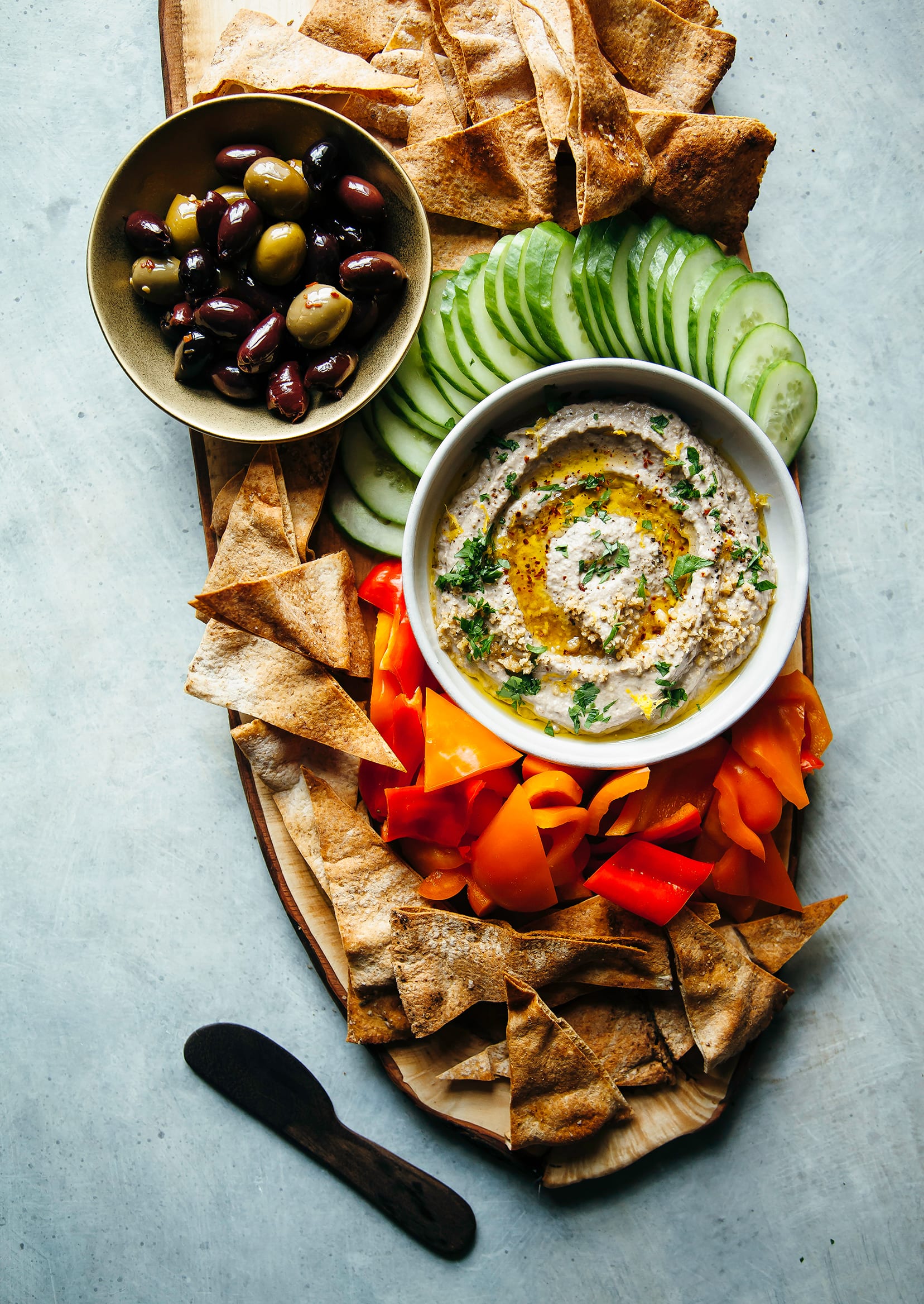 Image shows a board with a bowl of smooth eggplant dip, chopped vegetables, pita chips and olives.