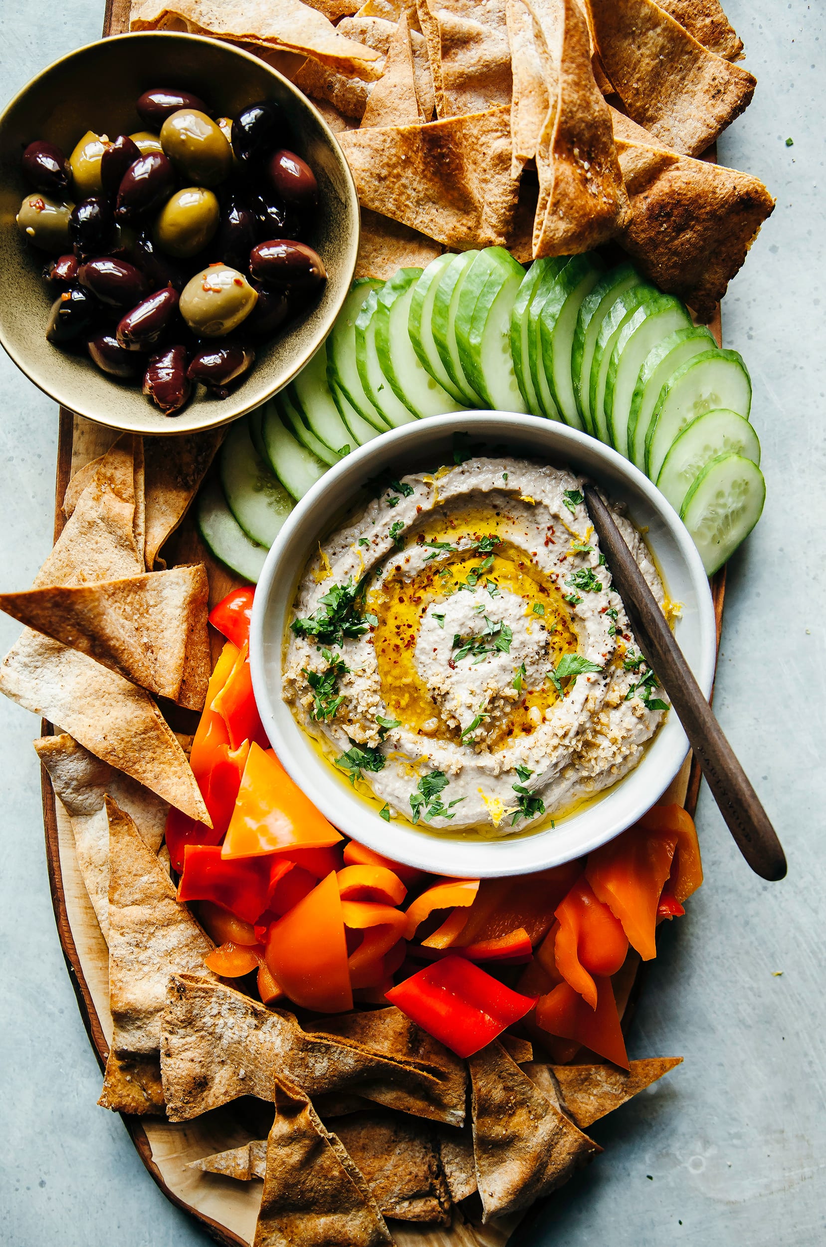 Image shows a board with a bowl of smooth eggplant dip, chopped vegetables, pita chips and olives.