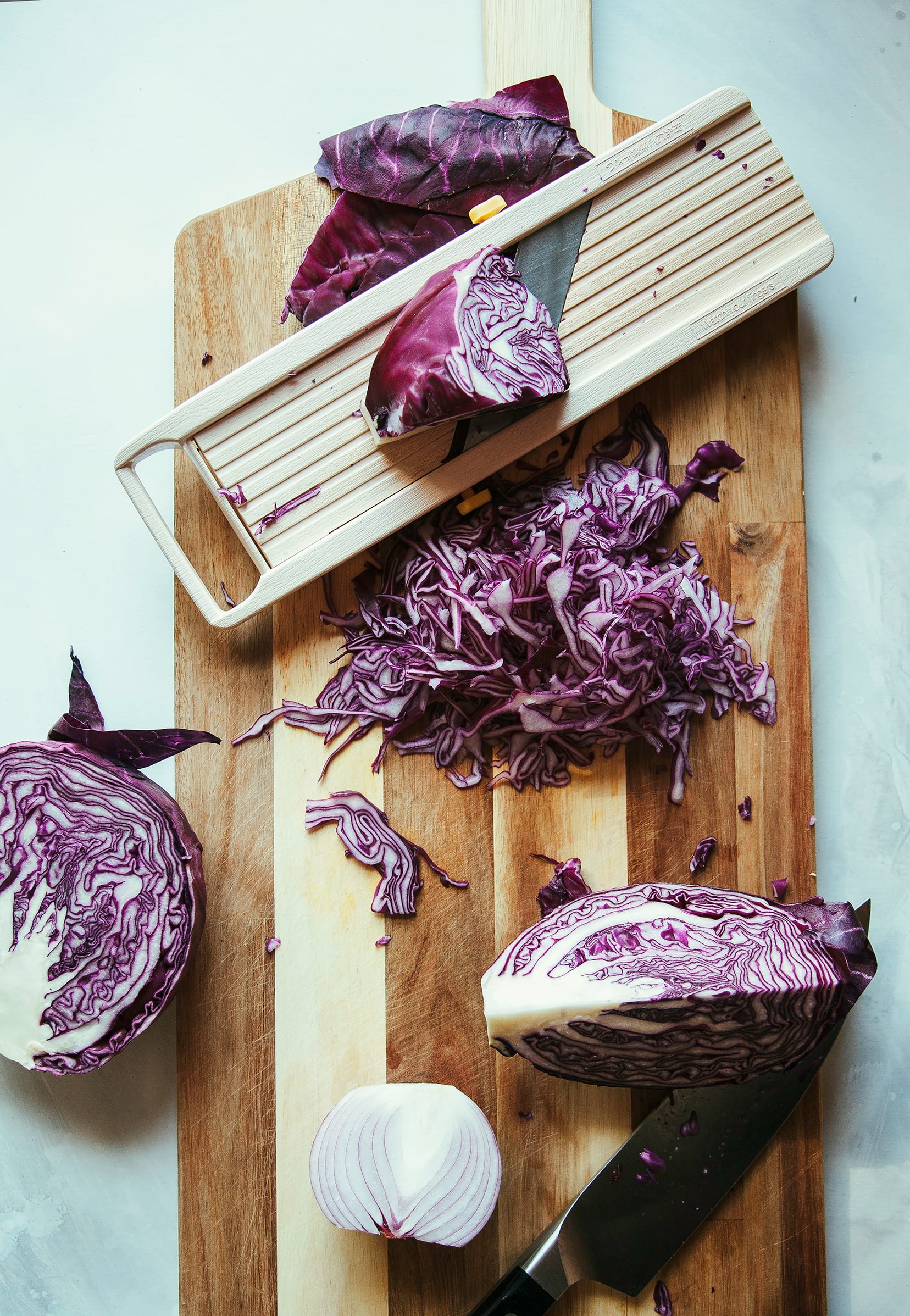 Overhead image of red cabbage being shredded with a mandolin over a wood cutting board.