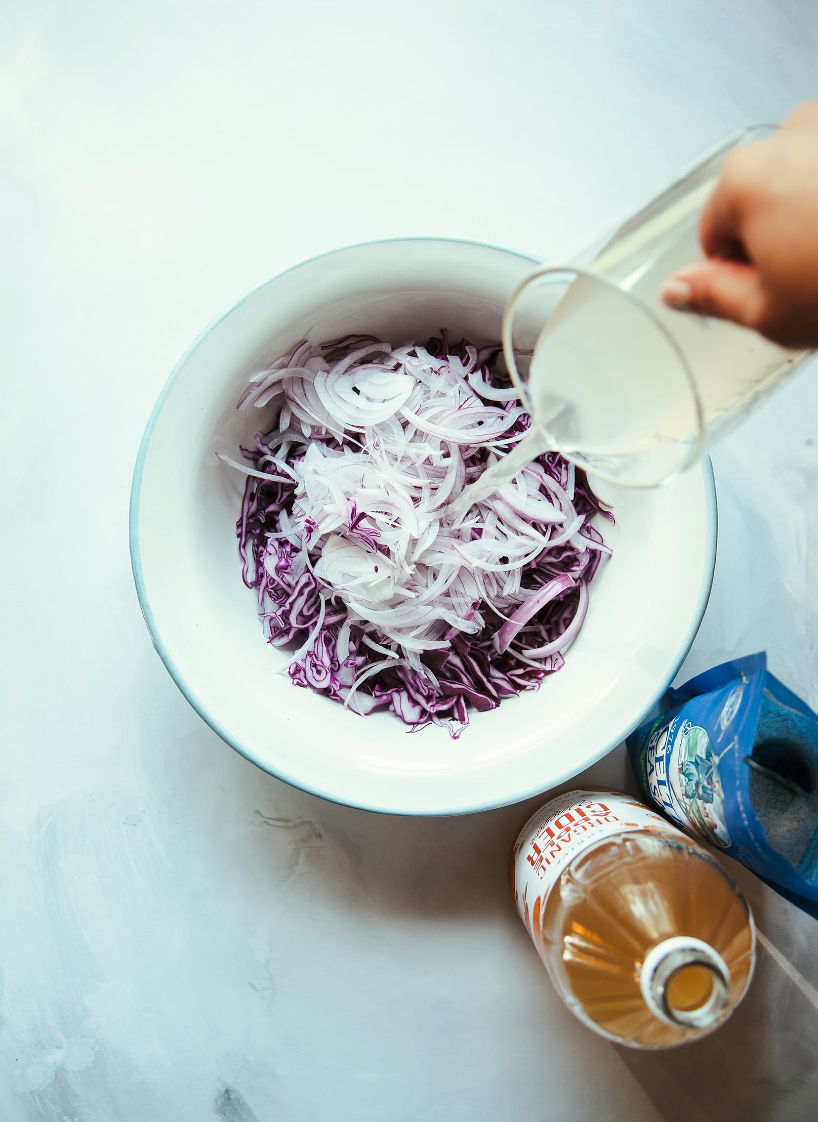 Image shows a hand pouring a vinegar mixture over a bowl of shredded red cabbage and onions.
