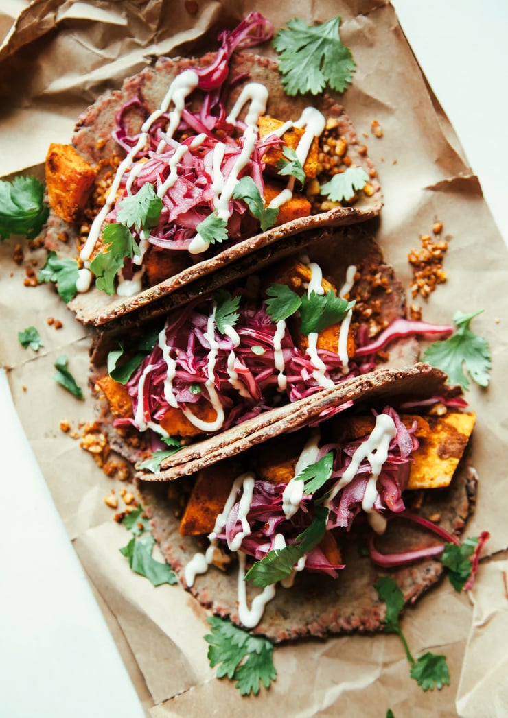 An overhead shot of 3 tacos containing a roasted sweet potato filling with pickled red cabbage on top. The tacos are seen on a crumpled piece of brown paper and are garnished with chopped herbs and a drizzles of a creamy white sauce.