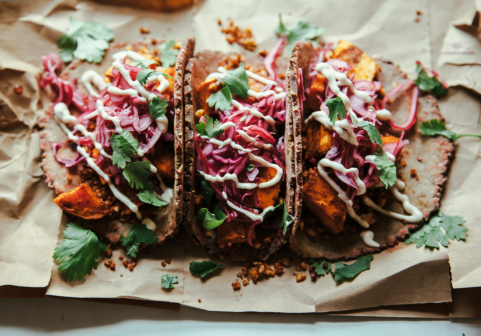 A 3/4 angle shot of 3 sweet potato tacos drizzled with a creamy white sauce and topped with chopped cilantro.