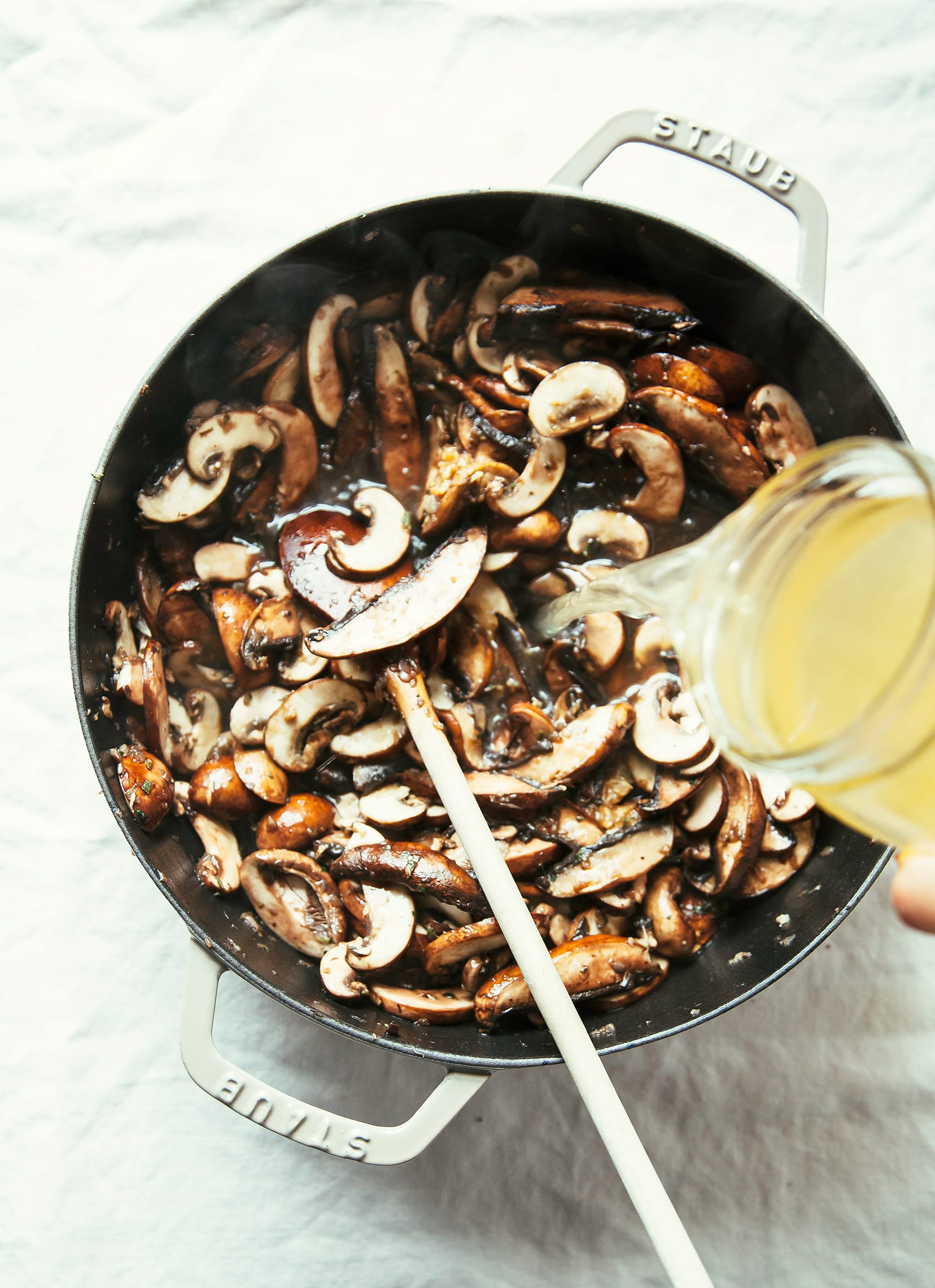 A hand is shown pouring vegetable stock into a pot of cooking mushrooms.