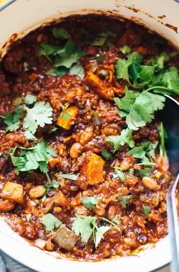 An up close, overhead shot of smoky squash chili with pinto and black beans. The chili is photographed in a white Dutch oven and is deep red in colour. The chili is garnished with bright green chopped cilantro.