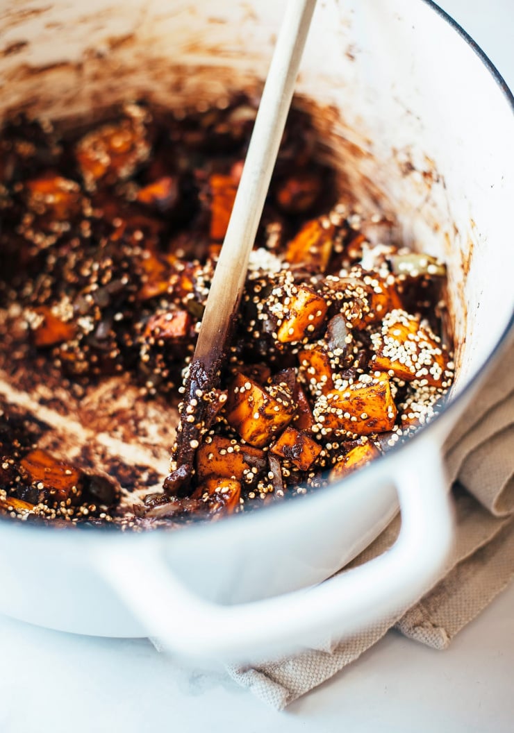 A spoon is stirring quinoa into a mix of squash and adobo sauce in a pot.