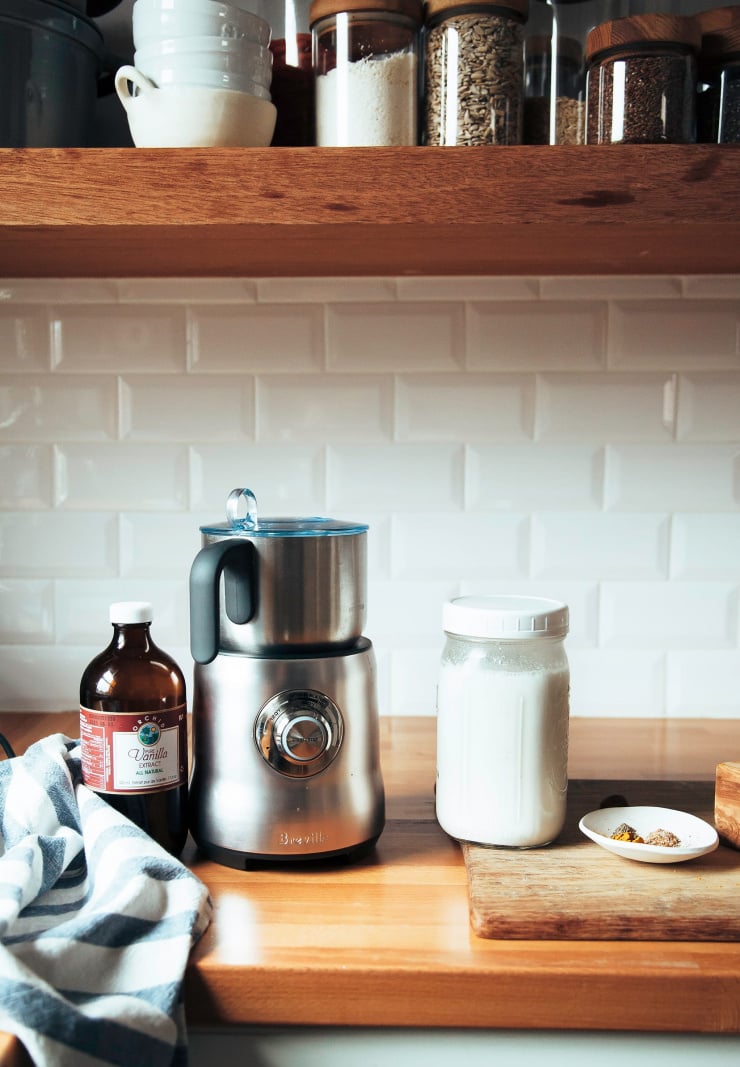 Image shows a milk frother on a kitchen counter with a jar of milk and a bottle of vanilla extract nearby.
