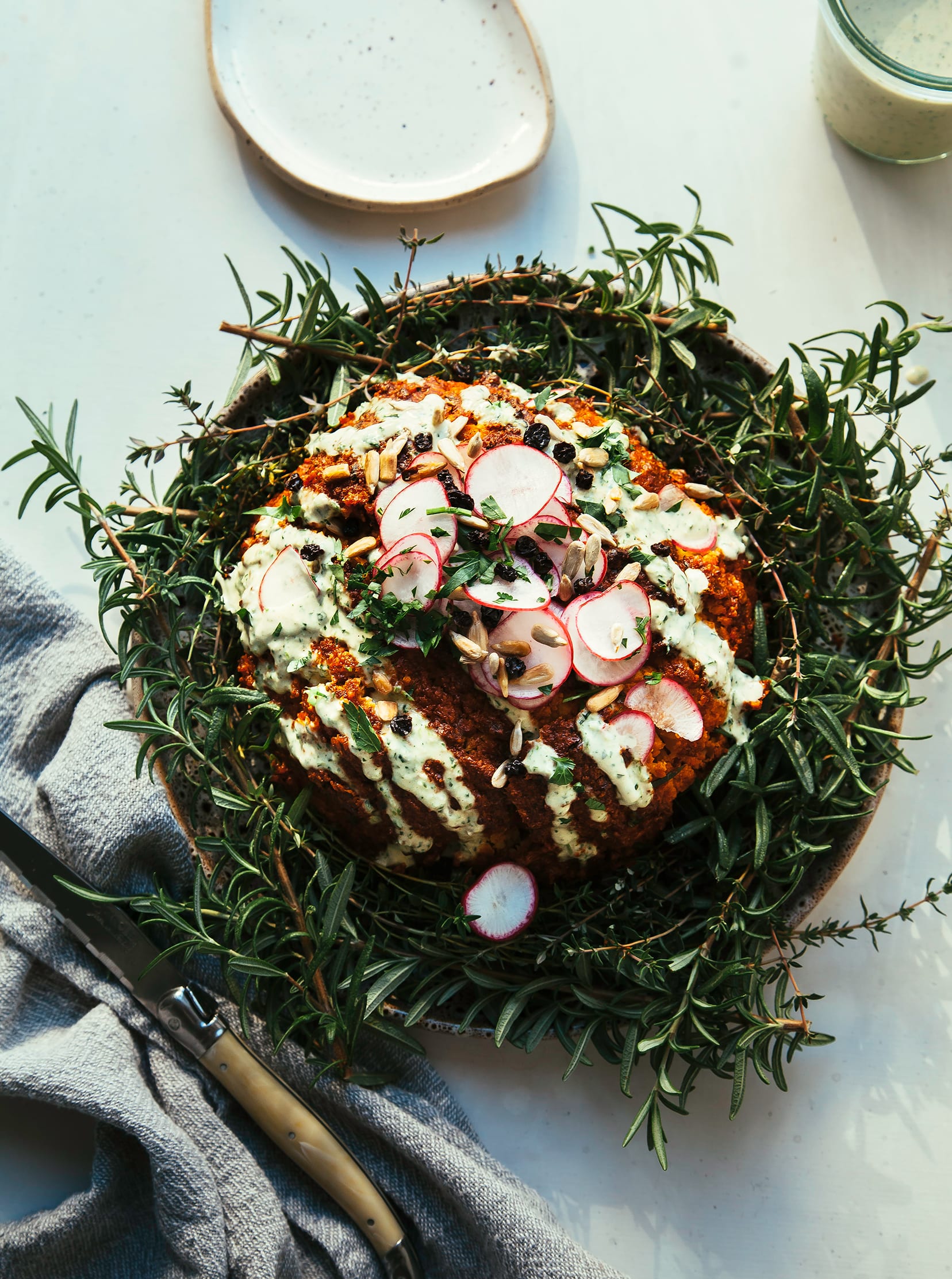 Image shows a whole roasted cauliflower coated in deep red romesco sauce. The cauliflower is garnished with a creamy light green sauce and sliced radishes.