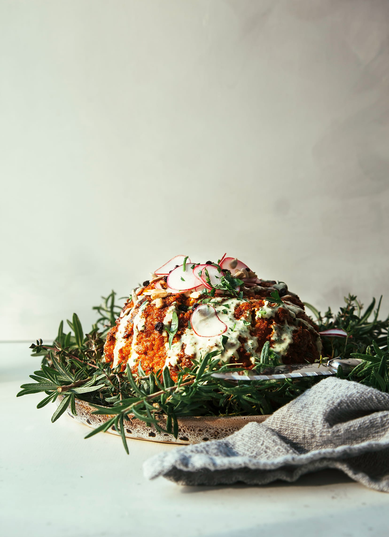 Image shows a whole roasted cauliflower coated in deep red romesco sauce. The cauliflower is garnished with a creamy light green sauce and sliced radishes.