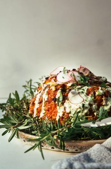 A head on shot of a whole roasted cauliflower coated in a res sauce and drizzled with a creamy, light green sauce. The platter is set with whole sprigs of herbs.