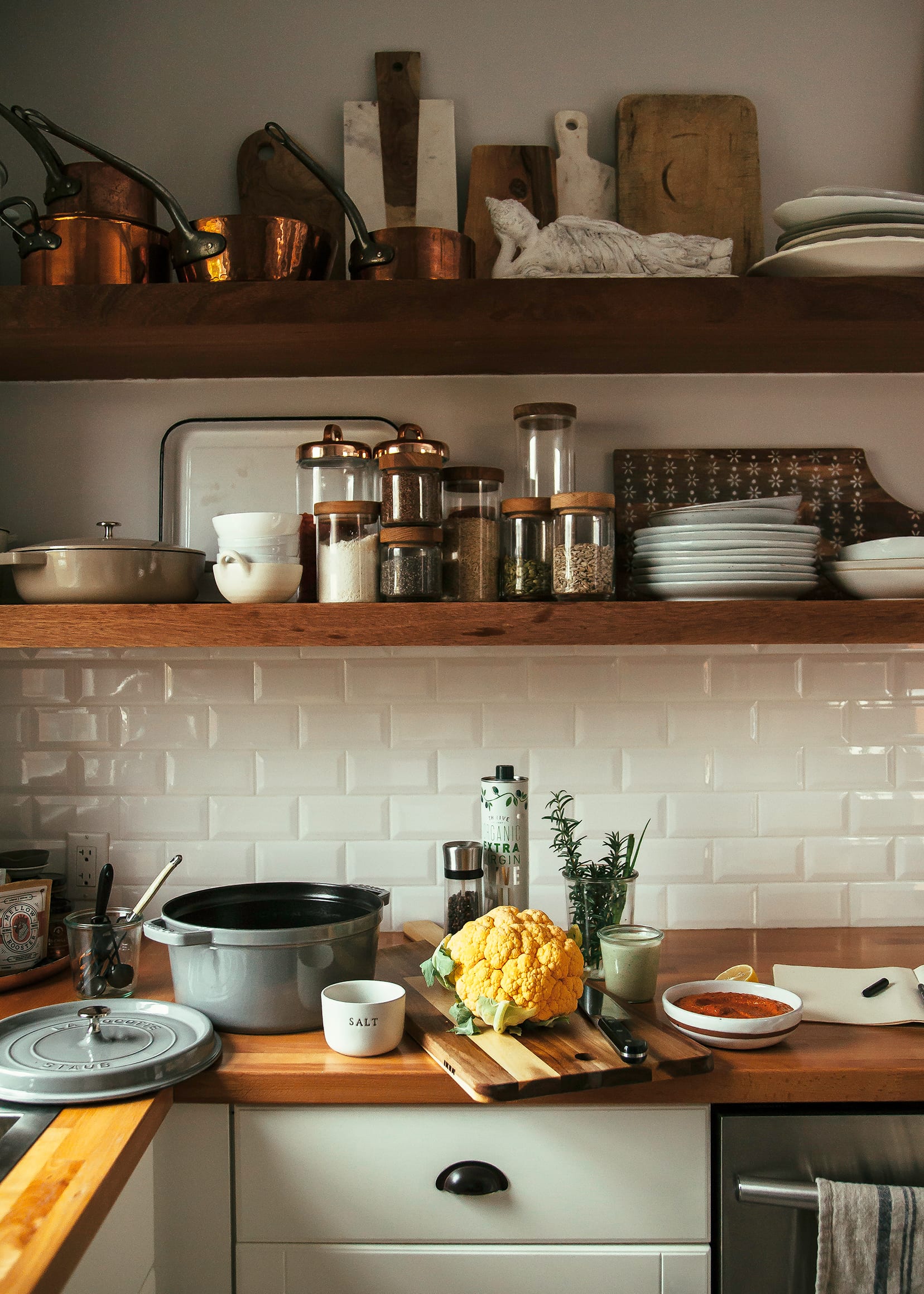 A kitchen scene shows a cauliflower and other prepped ingredients on top of a butcher block counter with open shelving in the background.