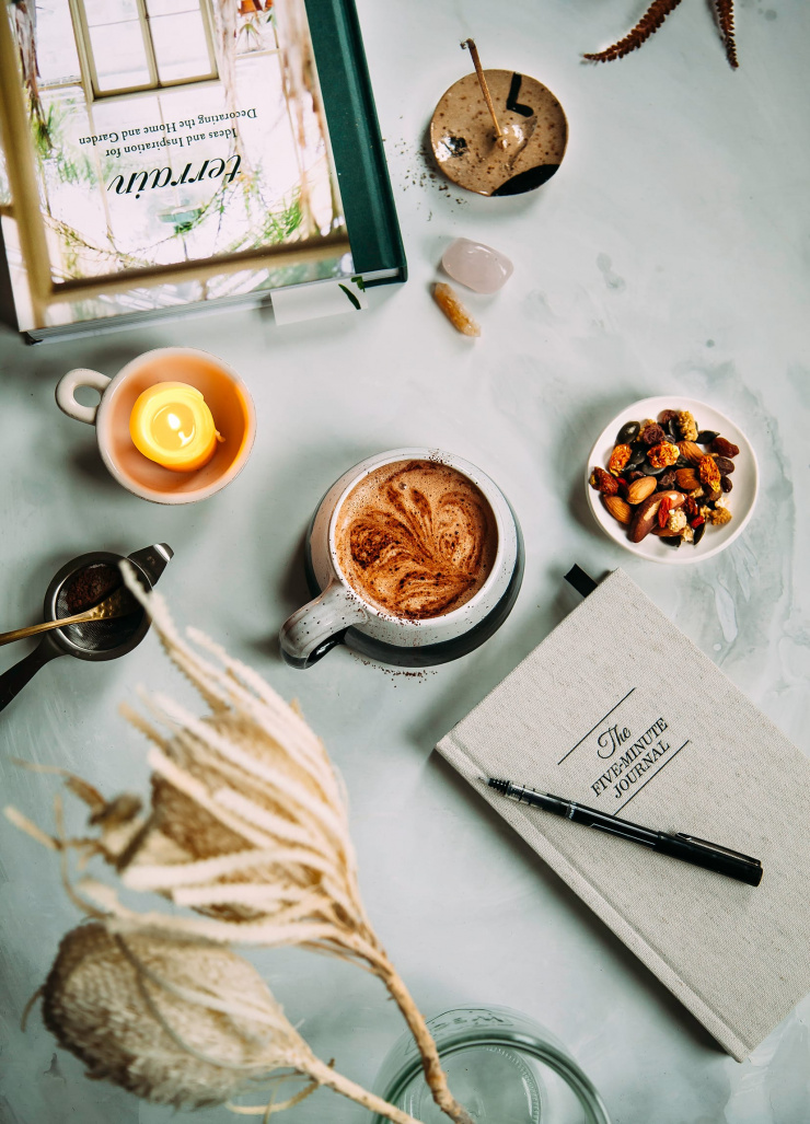 Overhead image shows a cup of hot cocoa, a lit candle, a book, a notebook, and a tray of trail mix.