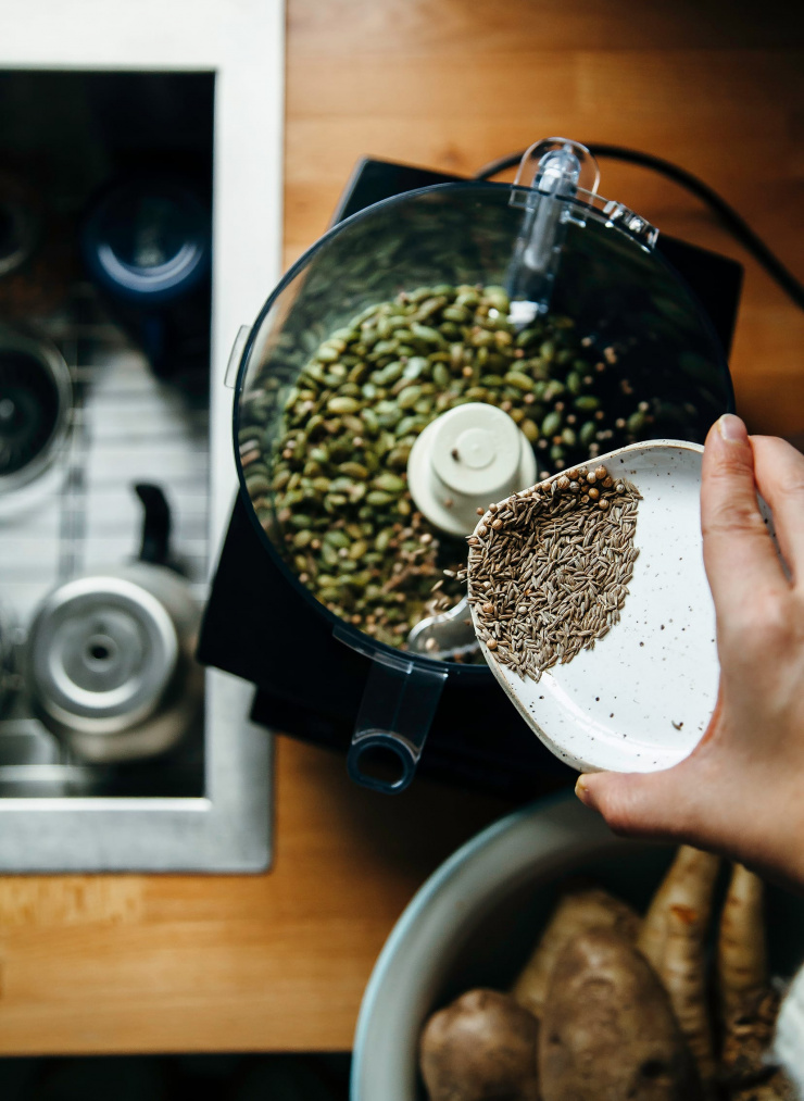 Image shows a hand pouring whole spices into a food processor.