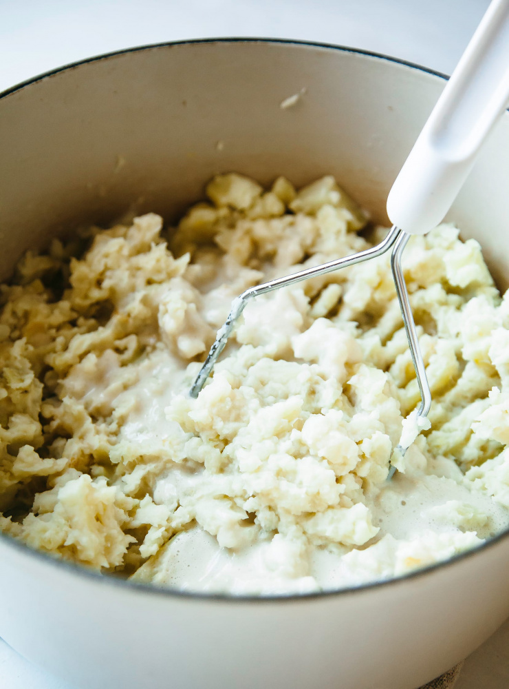 Image shows cooked root vegetables being mashed with a hand masher.