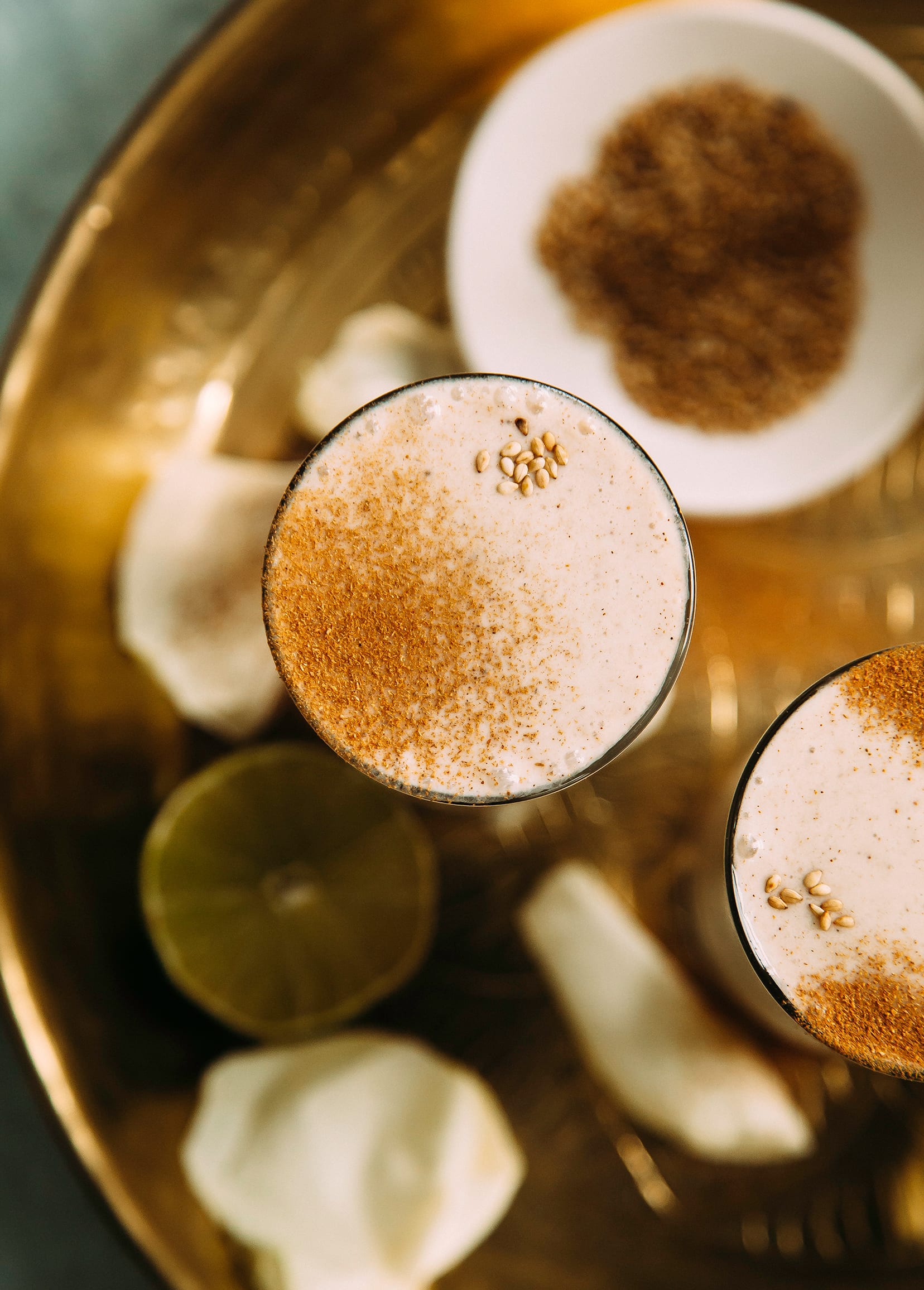 An overhead and up close shot of a creamy beige beverage in a slim clear glass beverage on top of a gold, etched tray. The beverage is garnished with ground cinnamon and sesame seeds.