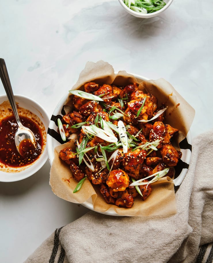 An overhead shot of sticky and crispy sesame cauliflower bites in a parchment paper-lined bowl. The bites are shiny with the deep red sauce on top.