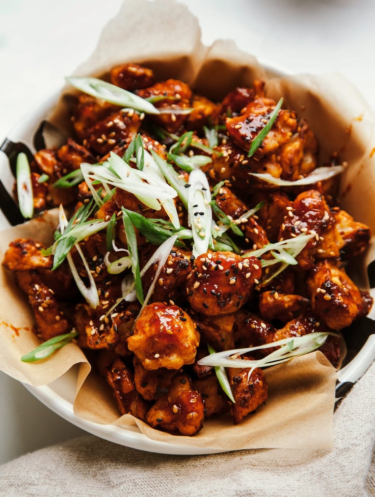 An overhead shot of sticky and crispy sesame cauliflower bites in a parchment paper-lined bowl. The bites are shiny with the deep red sauce on top.