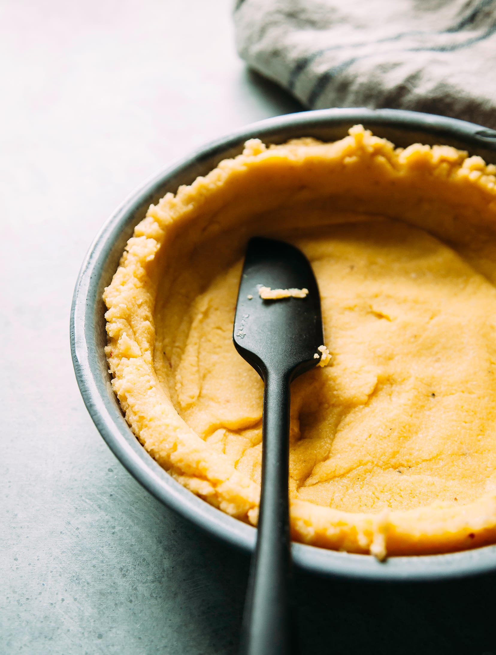 Image shows a spatula pressing a polenta-based crust into a circular baking dish.