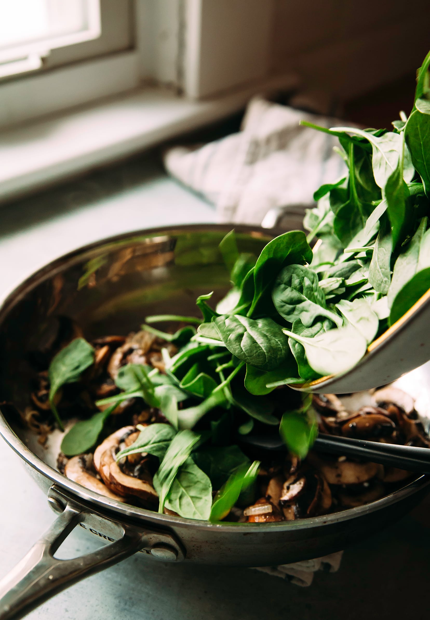 Image shows baby spinach being added to a sauté pan with mushrooms.