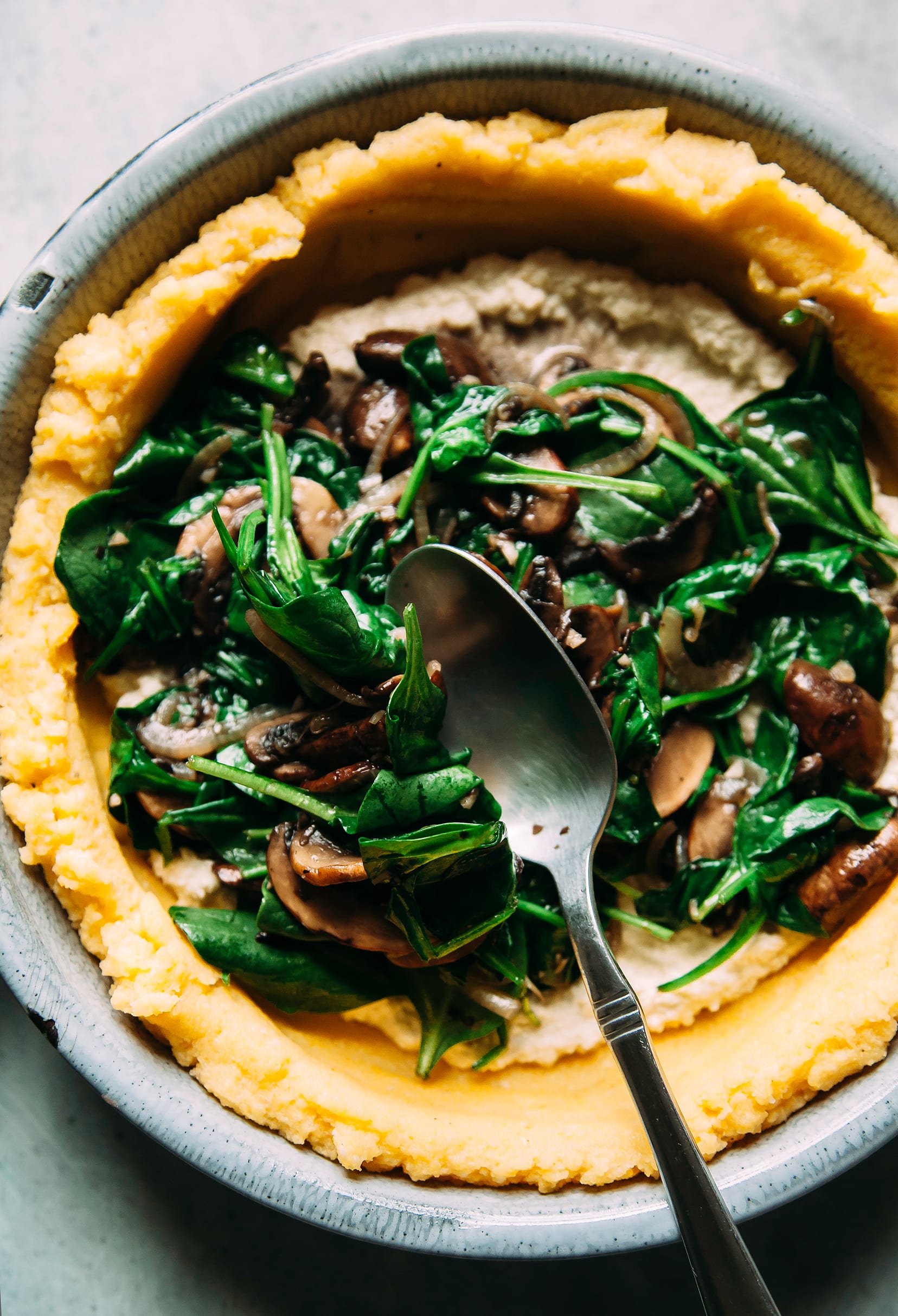 Image shows a spoon transferring a spinach and mushroom filling to a yellow polenta-based crust.
