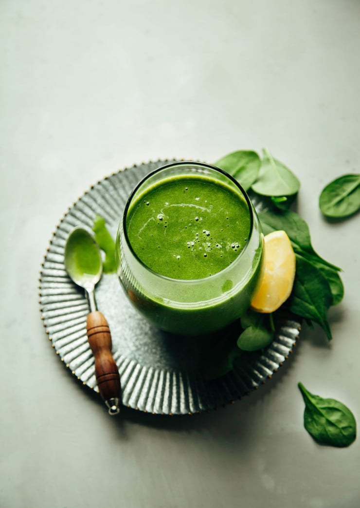 An overhead shot of a bright green smoothie in a clear, stemless wine glass thatโs perched on top of a galvanized metal plate on a grey background.