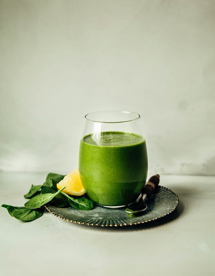 An head-on shot of a spinach banana & ginger smoothie in a clear, stemless wine glass thatโs perched on top of a galvanized metal plate on a grey background.