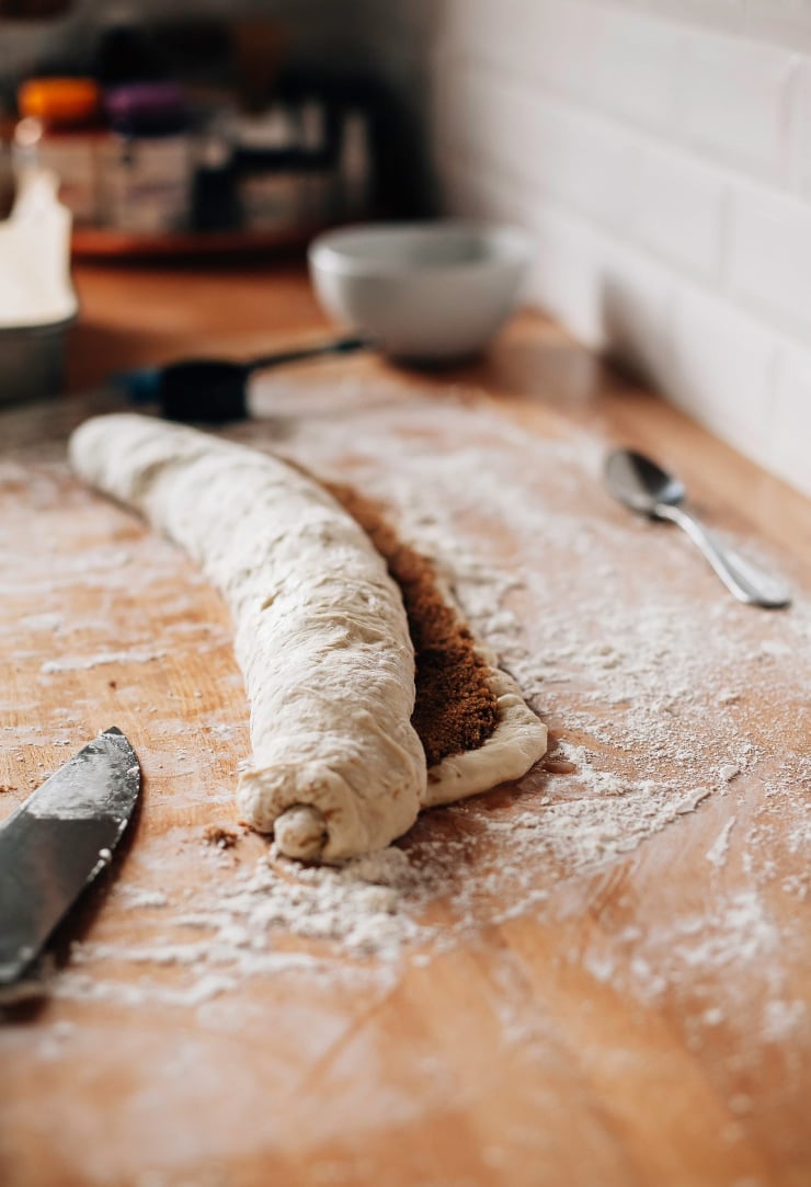 Image shows a rolled up tube of dough with a brown sugar filling on top of a butcher block countertop.