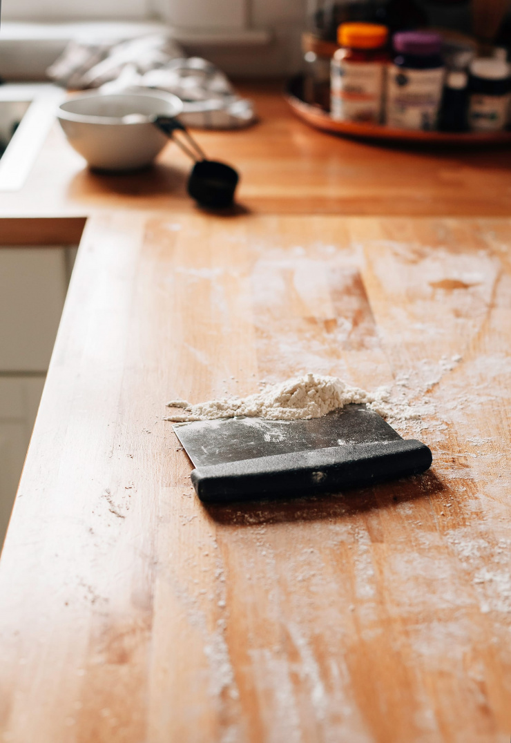 Image shows a bench scraper with gathered up dusting flour on top of a butcher block countertop.
