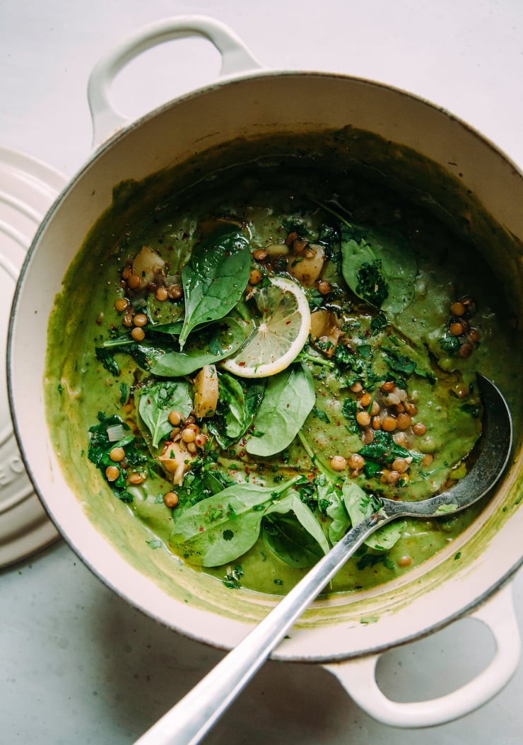 An overhead shot of deep green lentil stew in a white Dutch oven-style pot on a white background. The stew is bright green and garnished with extra baby spinach, cooked lentils, a slice of lemon, chili flakes, and little cooked bits of potato. A metal ladle hangs out of the pot.