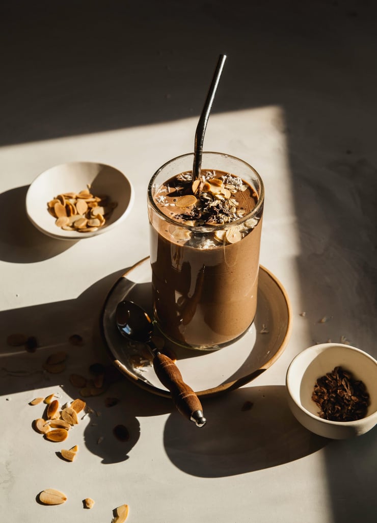 A 3/4 shot of a hearty chocolate oat smoothie in harsh light. The smoothie is in a clear glass on top of a white plate thatโs on top of a white background.