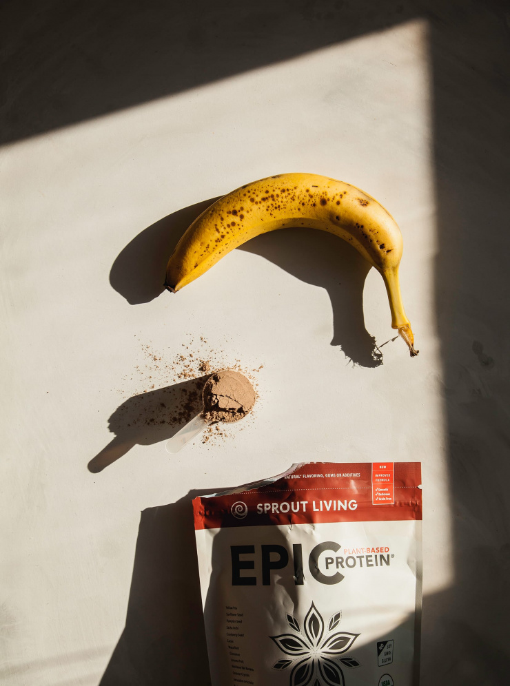 An overhead shot of a ripe banana and a scoop of protein powder in direct sunlight with strong shadows.