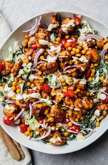 An overhead shot of a napa and kale slaw topped with chickpeas, crunchy sauce-coated cauliflower, red onion slices, and halved cherry tomatoes. All is coated in a creamy dressing and served in a wide white bowl.