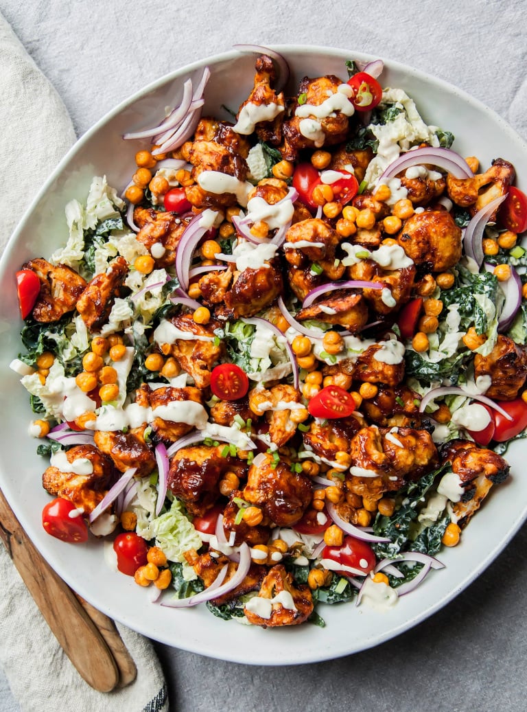 An overhead shot of a napa and kale slaw topped with chickpeas, crunchy sauce-coated cauliflower, red onion slices, and halved cherry tomatoes. All is coated in a creamy dressing and served in a wide white bowl.