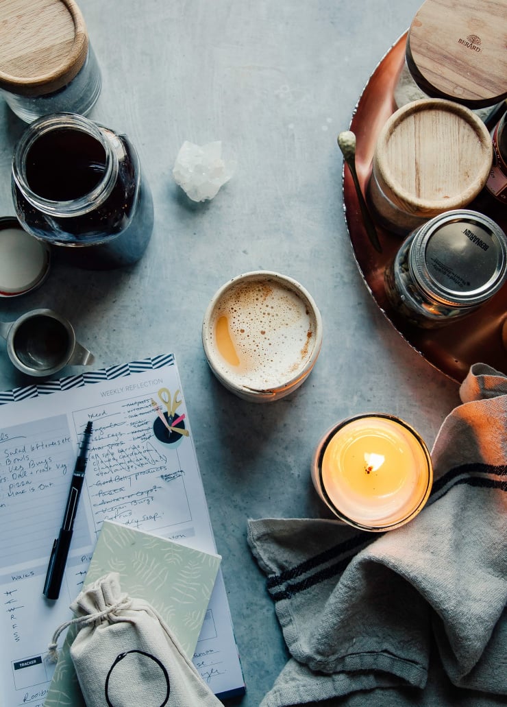 An overhead shot of a frothy tea latte next to a notebook and a bunch of jars and a lit candle.