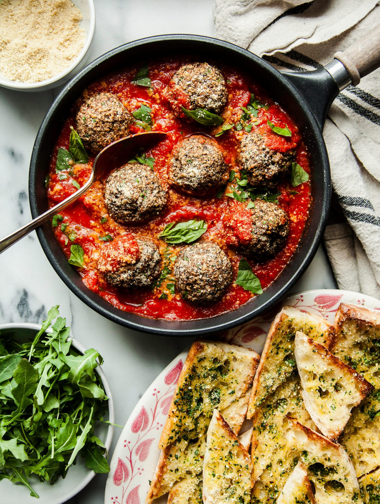 An overhead shot of vegan mushroom meatballs in a skillet, covered in marinara. A plate of garlic bread is shown to the side.