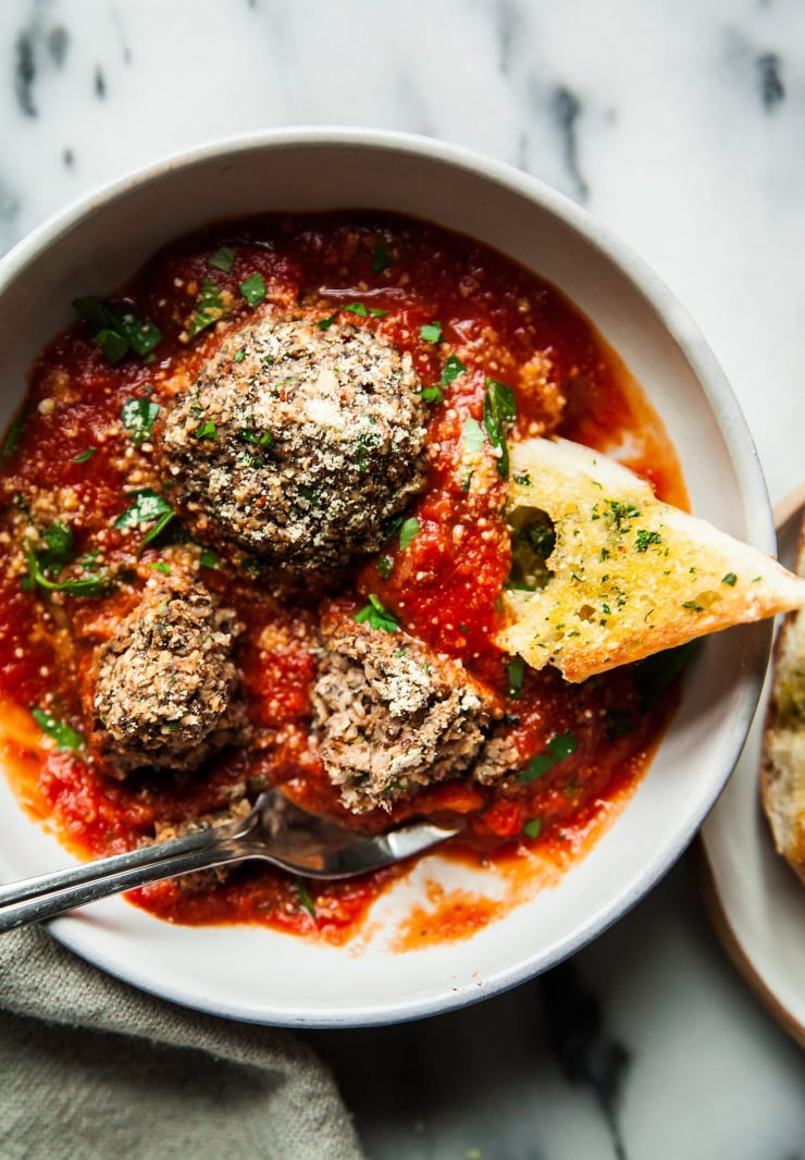 An overhead shot of vegan mushroom meatballs in marinara sauce in a bowl with a piece of garlic bread being dipped in the sauce.