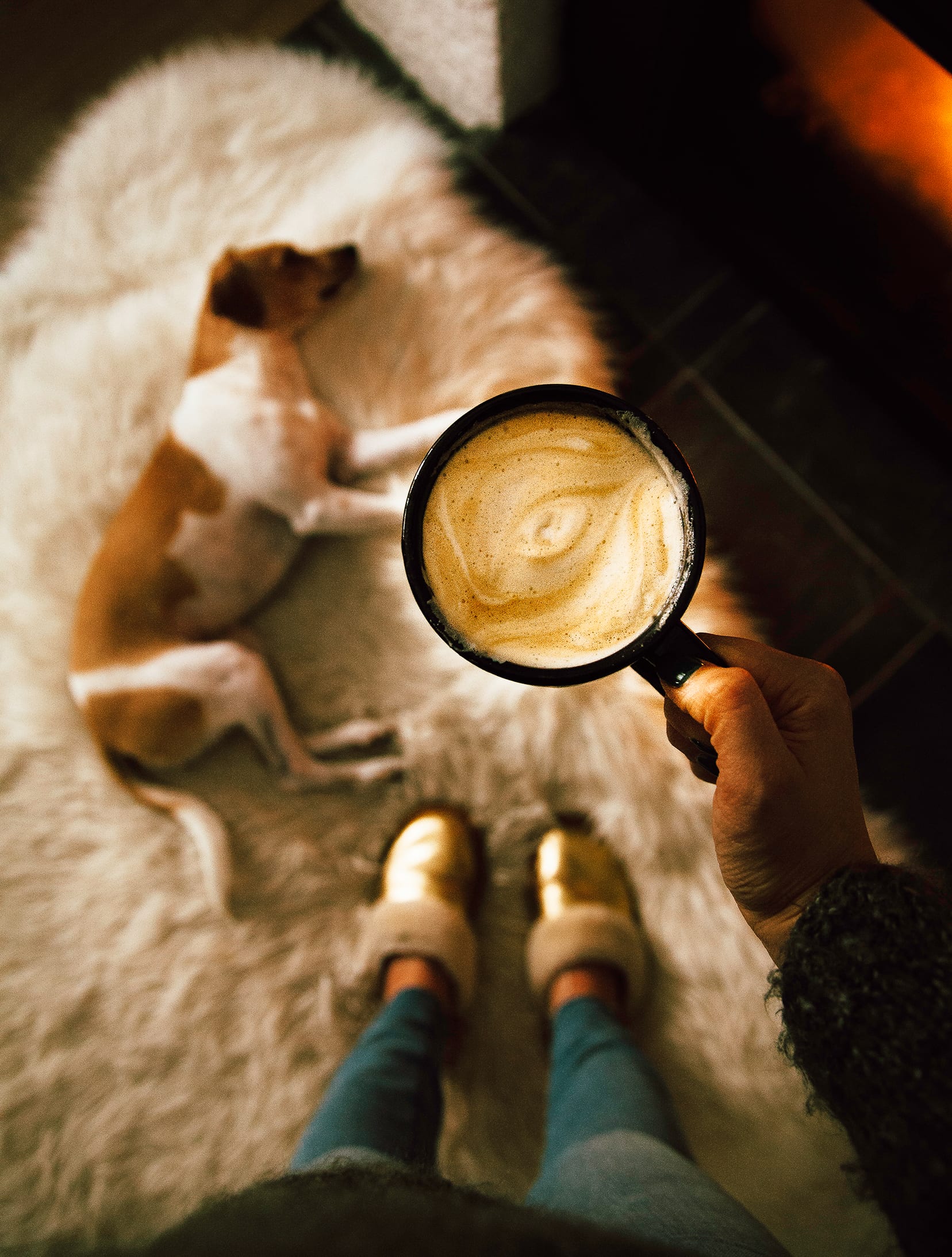 An overhead shot of a hand holding a mug of creamy coffee. A sleeping dog is in the background.