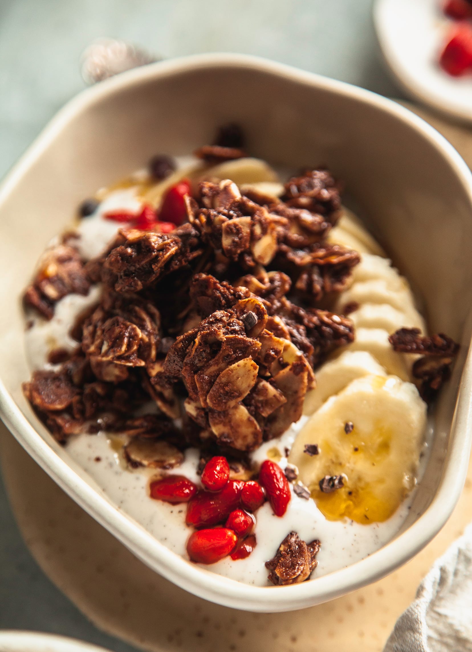 A 3/4 angle shot of a bowl of yogurt topped with deep brown granola, sliced banana and goji berries. Image is taken up close.