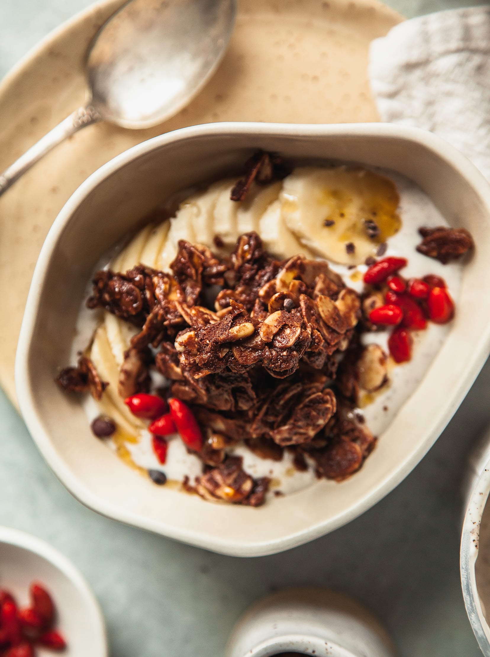 An overhead shot of a yogurt bowl with deep brown granola on top, plus goji berries and sliced banana. A tray of the granola is nearby.