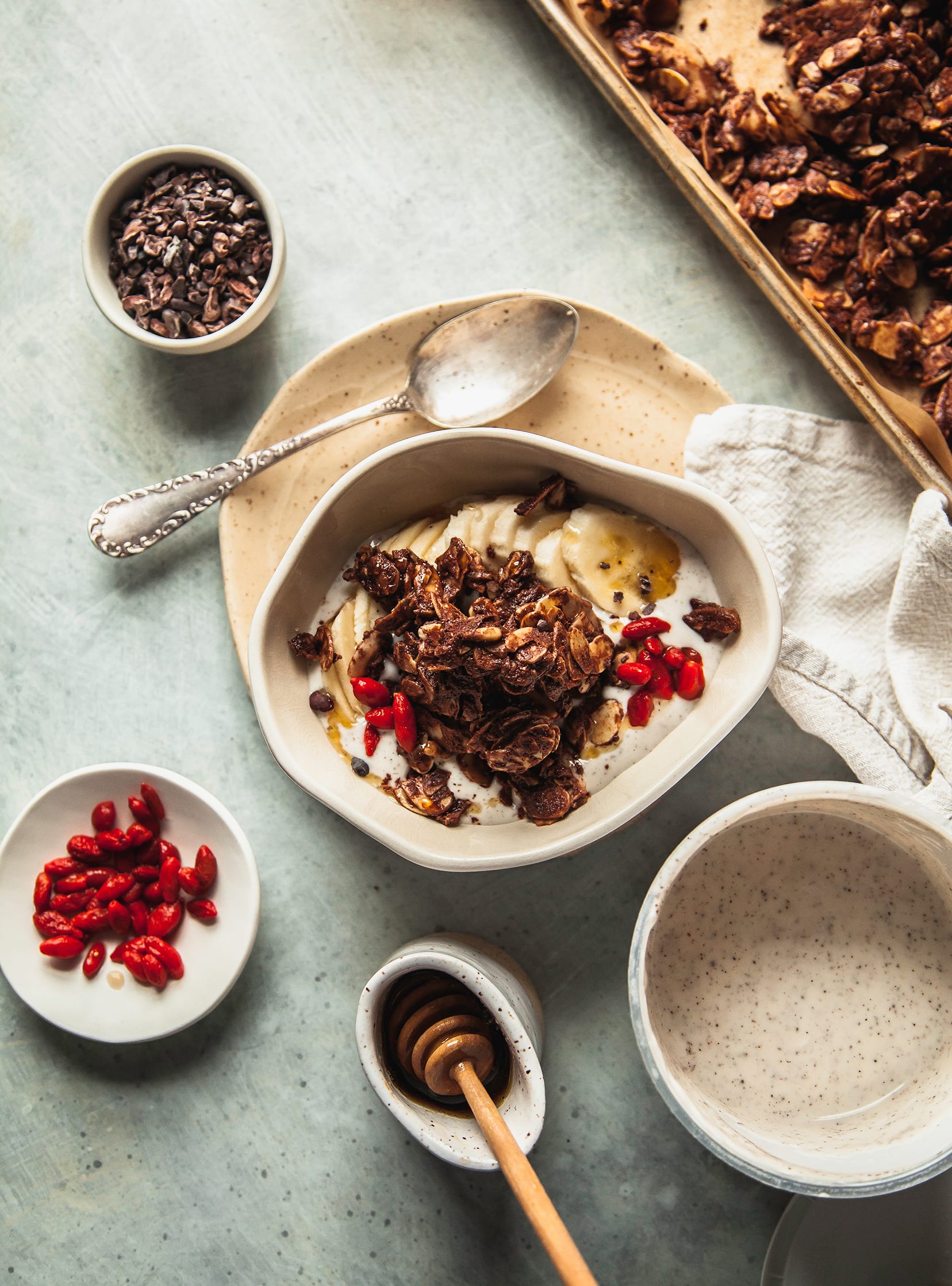 An overhead shot of a yogurt bowl with deep brown granola on top, plus goji berries and sliced banana. A tray of the granola is nearby.