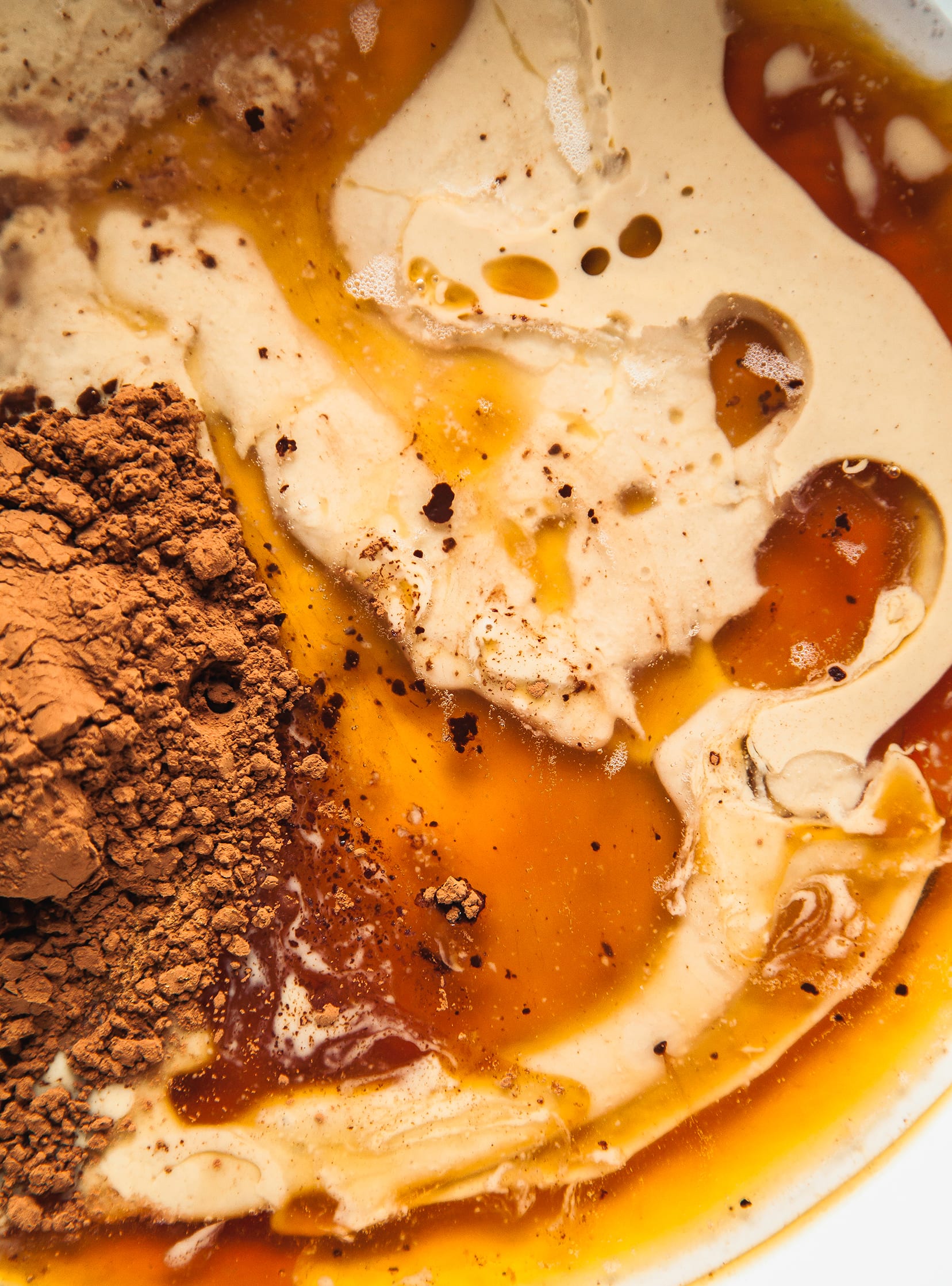 An up close, overhead shot of the wet ingredients for binding granola: tahini, cocoa, maple syrup, and vanilla all swirled together.