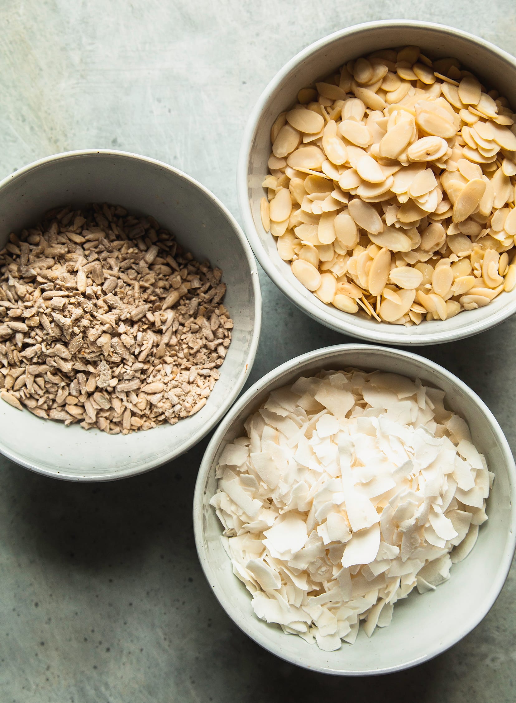 An overhead shot of ingredients in bowls: sunflower seeds, sliced almonds, and large coconut flakes.