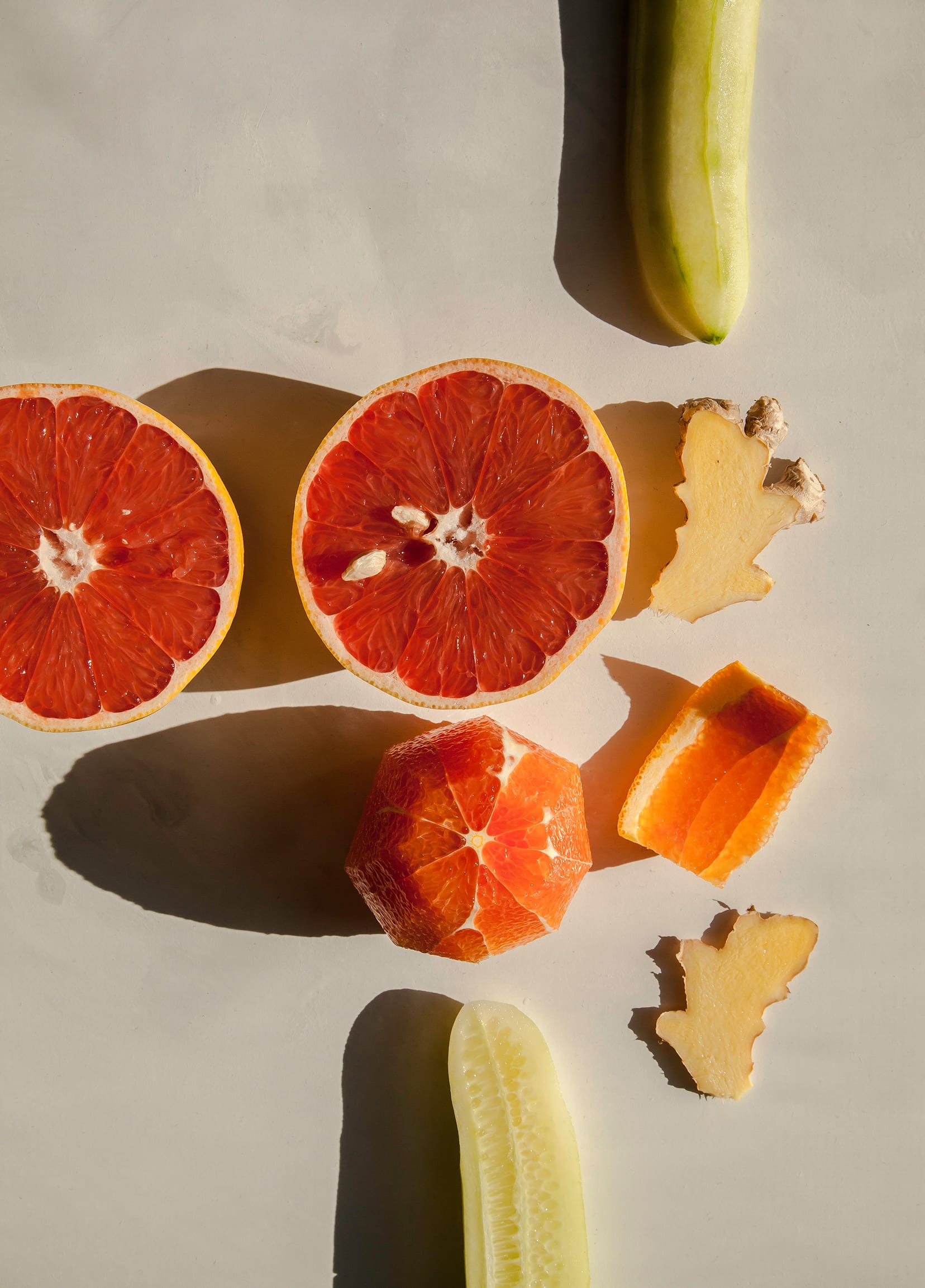 Over head shot of a halved grapefruit, peeled cucumber, orange, and sliced ginger in direct sunlight.