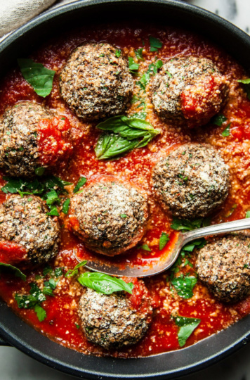 An overhead shot shows a pan of vegan mushroom meatballs swimming in marinara sauce. The meatballs are garnished with vegan parmesan and small basil leaves.