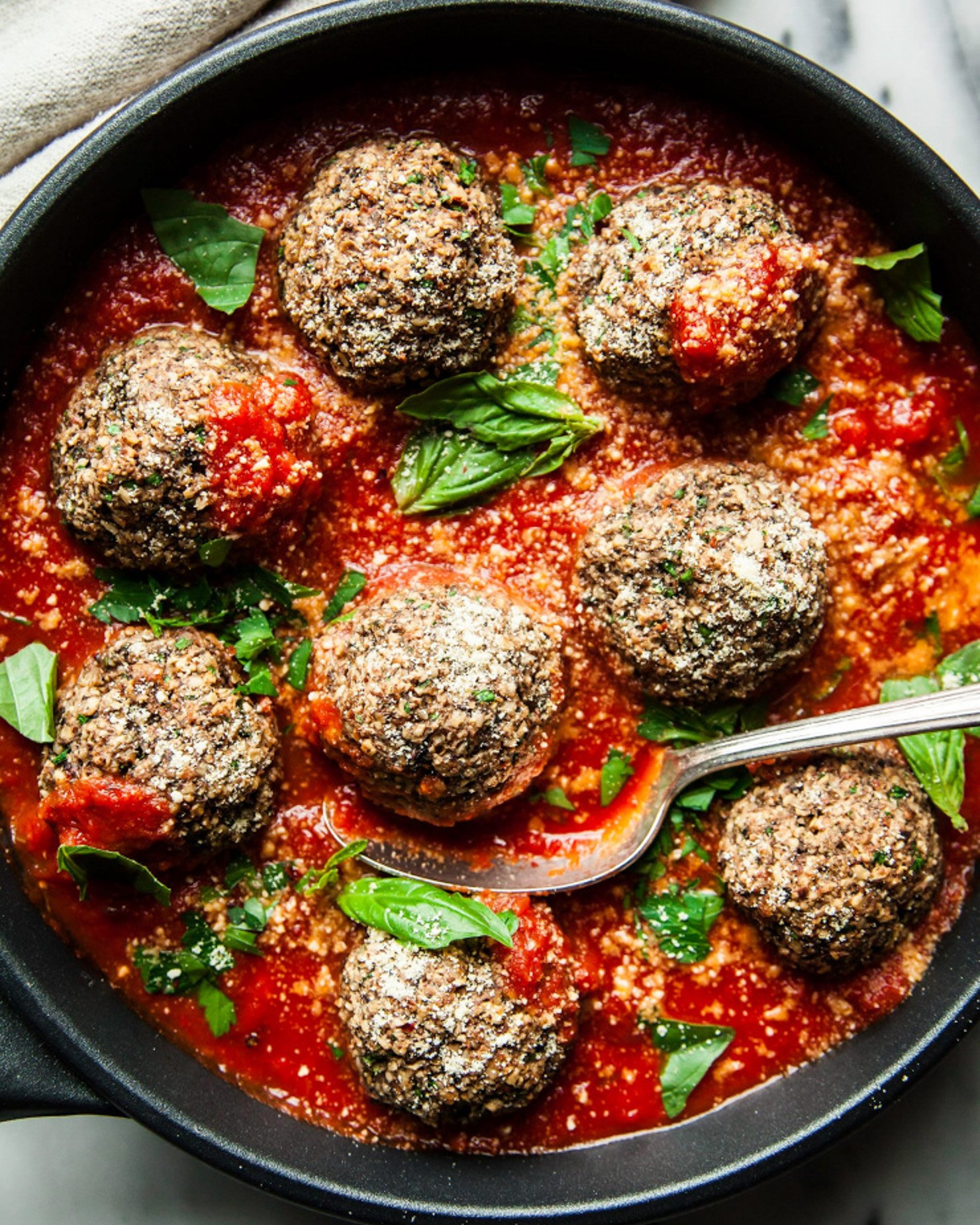 An overhead shot shows a pan of vegan mushroom meatballs swimming in marinara sauce. The meatballs are garnished with vegan parmesan and small basil leaves.