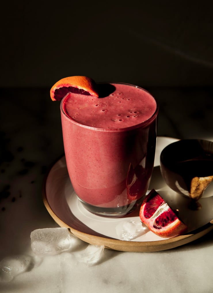 A 3/4 angle shot of a deep burgundy smoothie in a clear glass in harsh sunlight. A sliced blood orange is perched on a plate nearby. Included in a roundup of vegan smoothies.