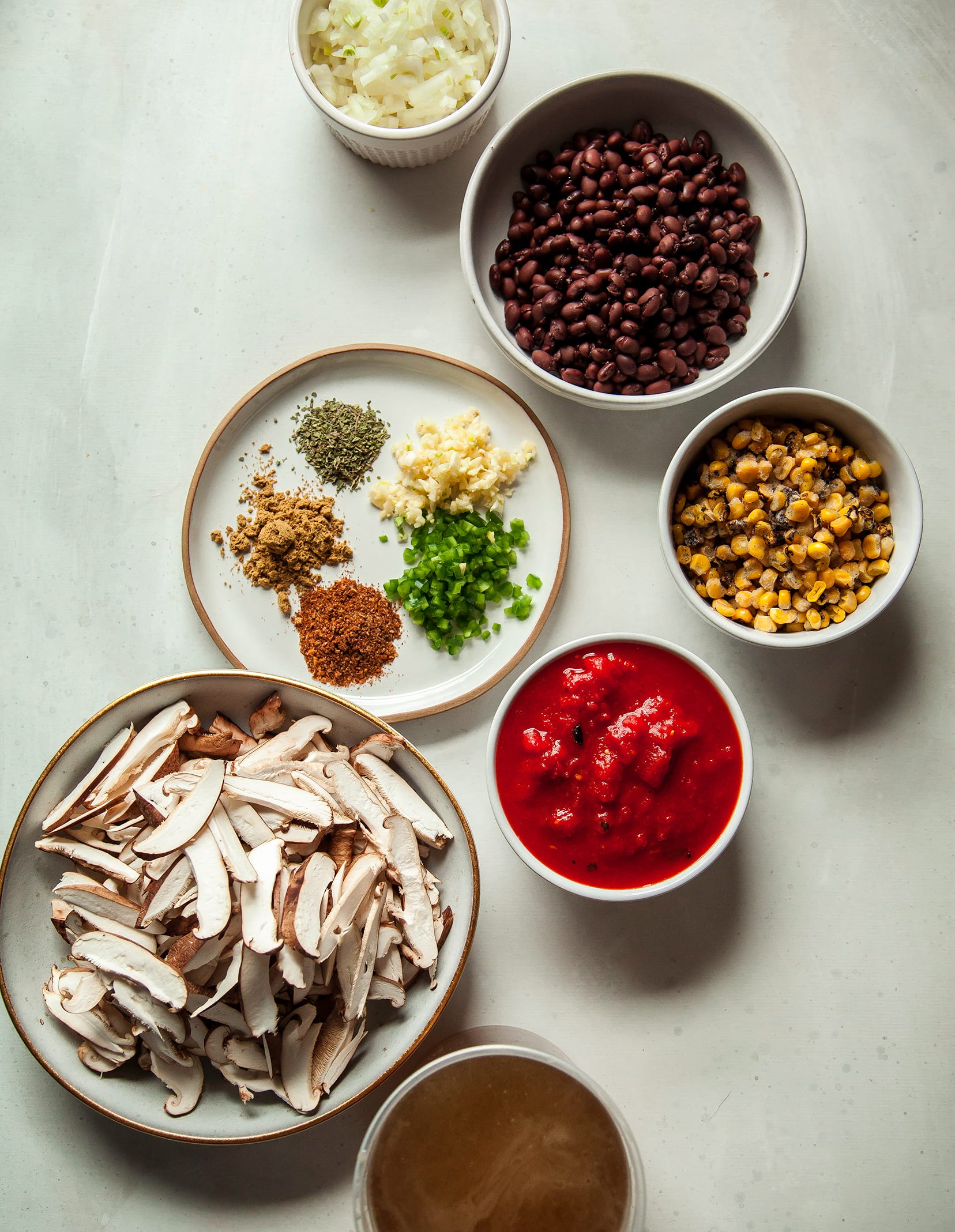 An overhead shot of ingredients, including: spices, garlic, jalapeño, canned crushed tomatoes, sliced shiitake mushrooms, black beans, corn, and diced onion. All on a white background.