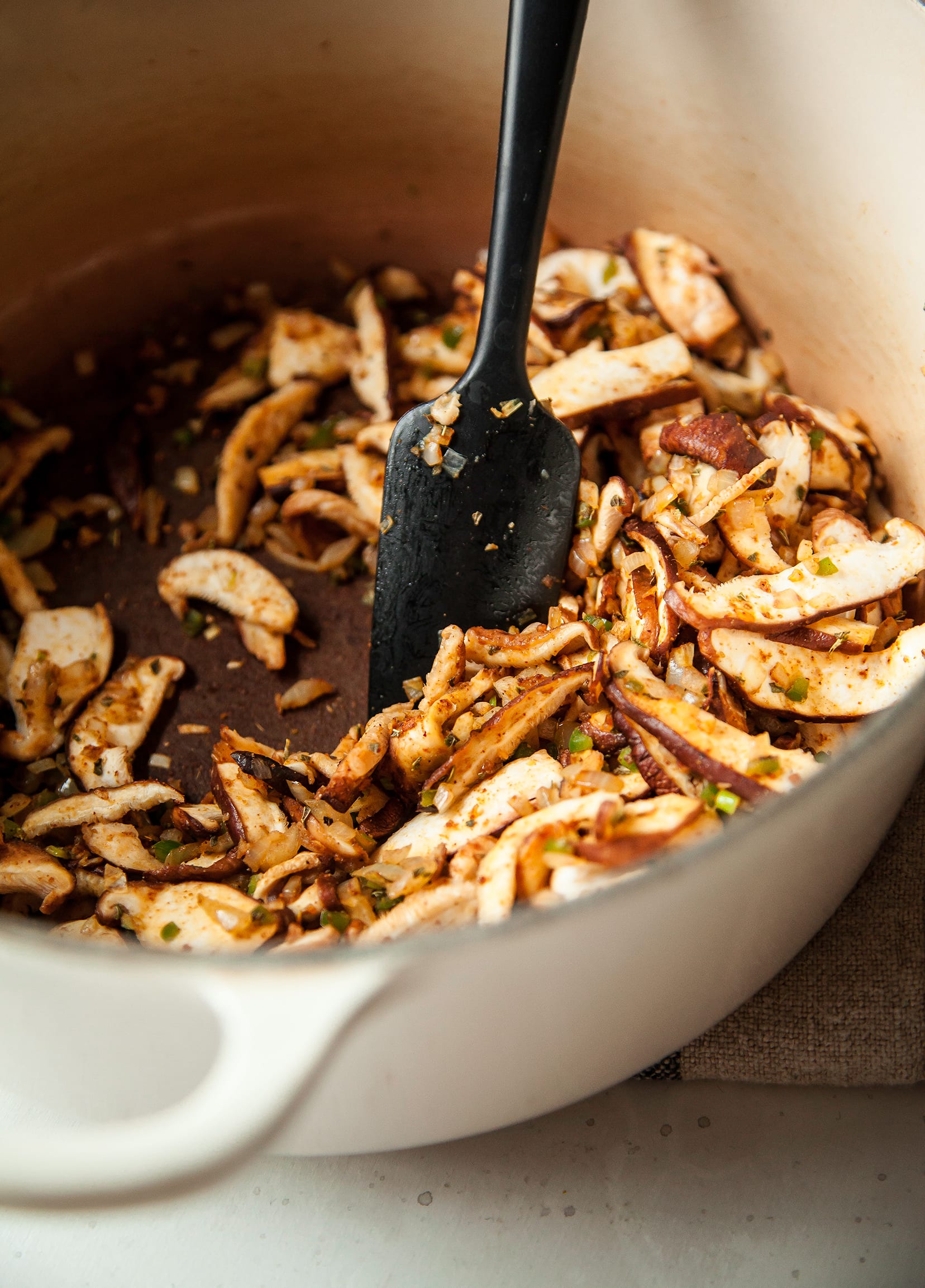 An up close shot of shiitake mushrooms being sautéed in a beige Dutch oven and stirred with a black spatula.