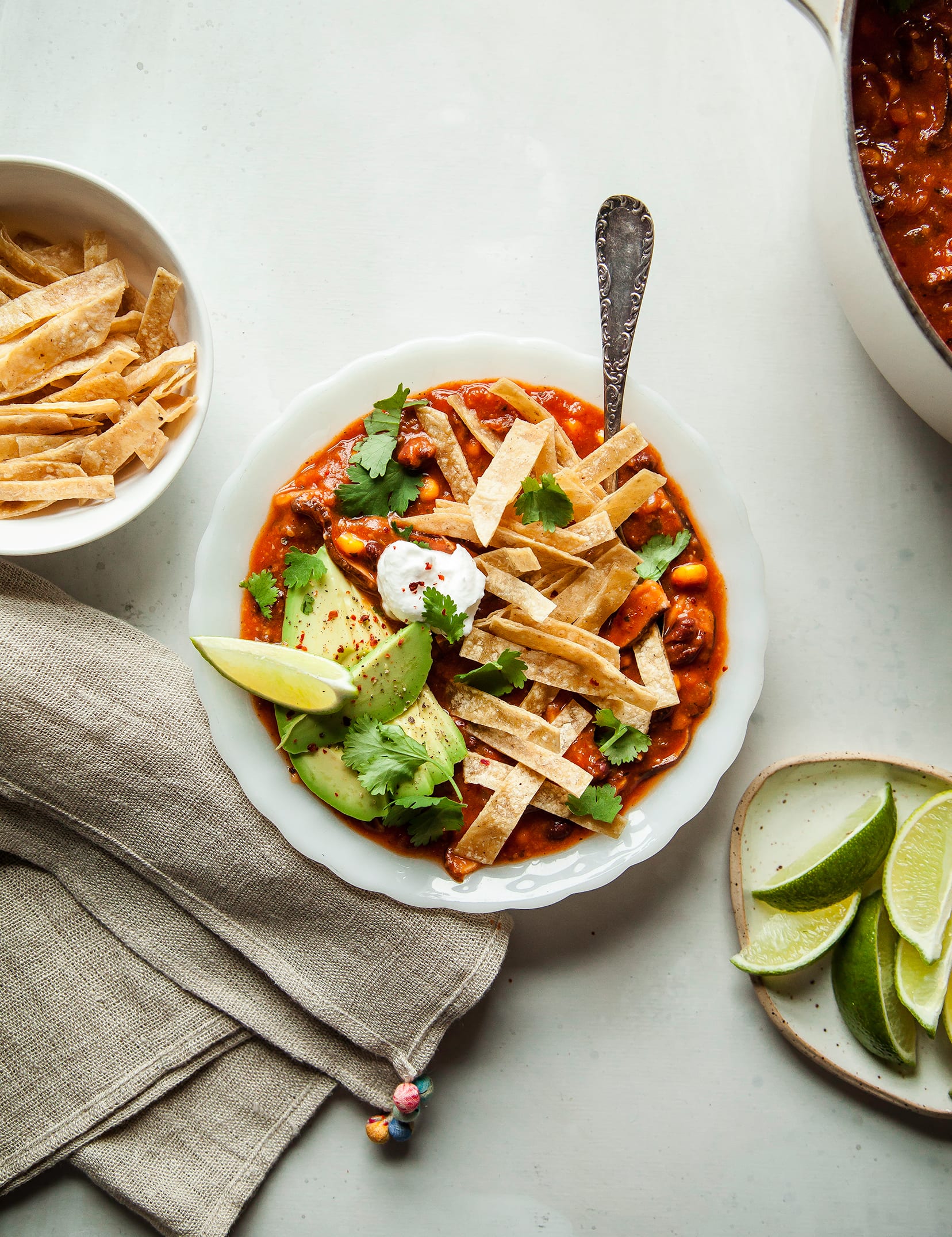 An overhead shot of smoky shiitake tortilla soup in a white bowl on a white background. The soup is topped with crispy tortilla strips, a lime wedge, vegan sour cream, and chopped cilantro.