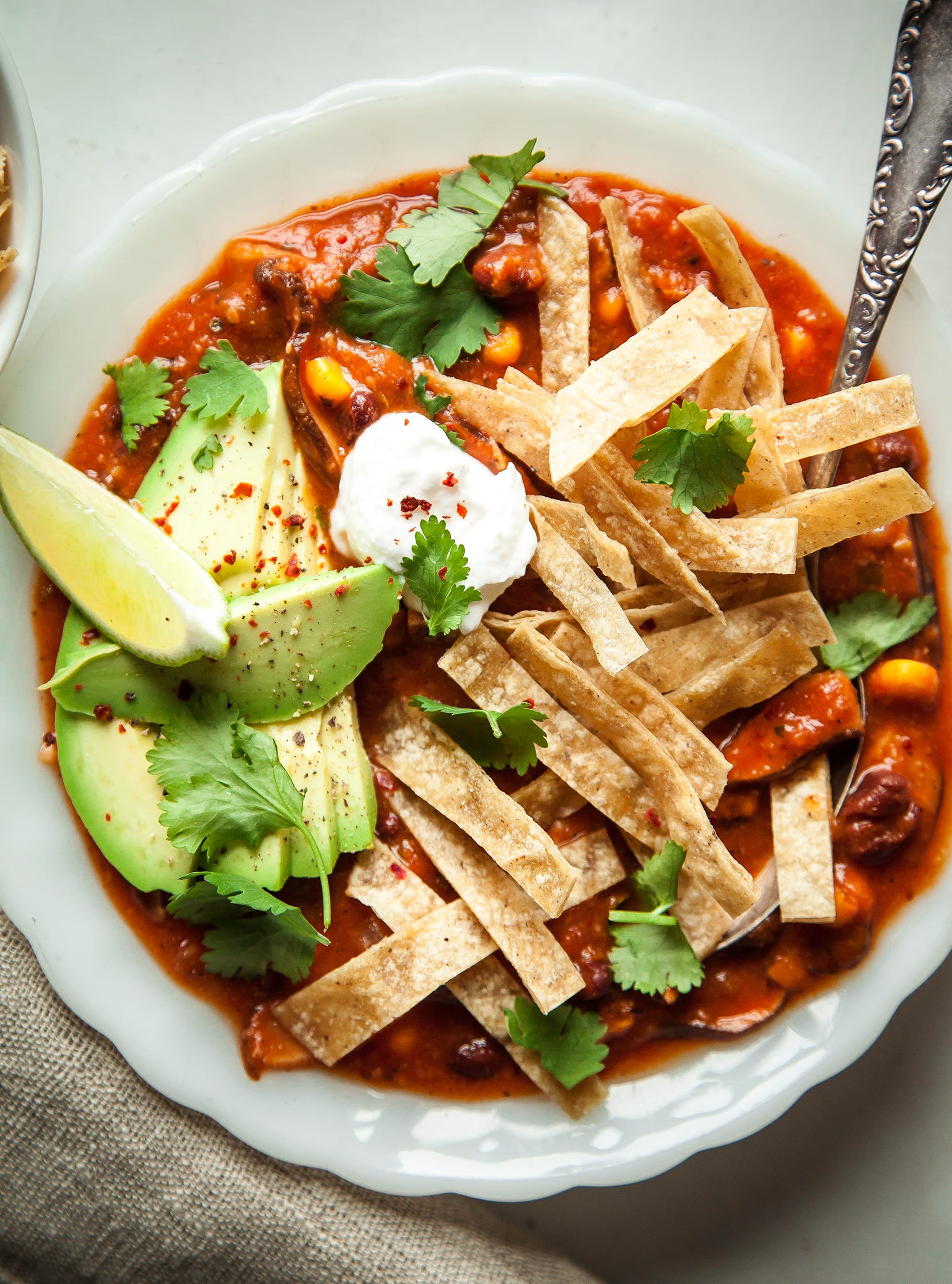 An overhead shot of smoky shiitake tortilla soup in a white bowl on a white background. The soup is topped with crispy tortilla strips, a lime wedge, vegan sour cream, and chopped cilantro.