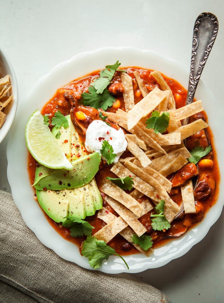 An overhead shot of smoky shiitake tortilla vegan soup recipe in a white bowl on top of a white background. The deep red soup is garnished with sliced avocado, a dollop of vegan sour cream, sliced avocado, tortilla strips, and fresh cilantro.